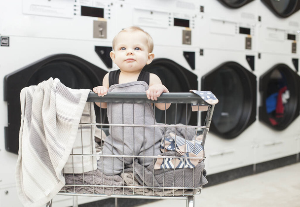 Portrait of funny baby boy wearing jumpsuit sitting in laundry - Foto, afbeelding