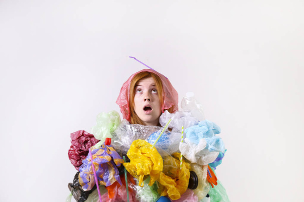 Emotional redhead woman looking stressed, covered with eco harmful plastic garbage, bags and straws of different color. Zero waste concept. Sad female frowning, angry about littering. - Zdjęcie, obraz