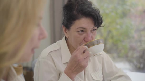 Mature Caucasian brunette woman drinking tea or coffee from the cup and talking. Blond senior lady listening her at the foreground. Friends or sisters resting together indoors. - Кадры, видео