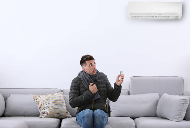Young man with air conditioner remote control freezing on sofa at home - Φωτογραφία, εικόνα