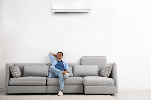 Happy young man relaxing on sofa under air conditioner at home - Foto, Imagen