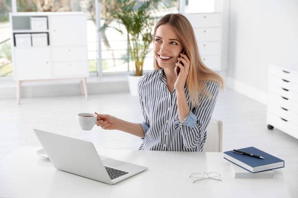 Young businesswoman talking on phone while using laptop at table in office - Foto, Imagen