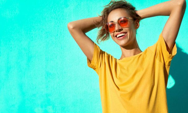 Fashionable cheerful young woman posing on blue background, wearing yellow t-short and trendy red eyeglasses. - Photo, Image