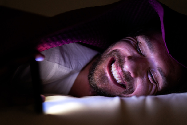 Bearded young man is lying in bed under his blanket. He cannot s - Foto, afbeelding