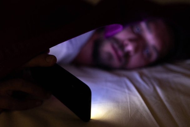 Bearded young man is lying in bed under his blanket. He cannot s - Foto, Imagen