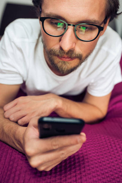 Rather serious young man wearing glasses lying in bed while look - Foto, Imagem