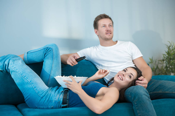 young cute couple beautiful brunette woman lying on handsome mans leg while watching movie on the sofa and eating popcorn sweet moments - Photo, Image
