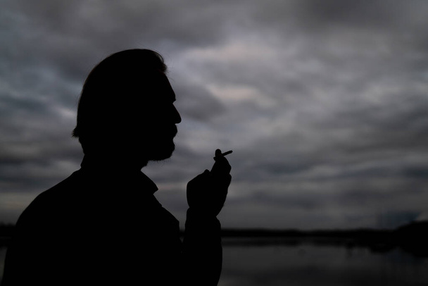 Dark silhouette of a handsome young man smoking a cigarette. Dar - 写真・画像