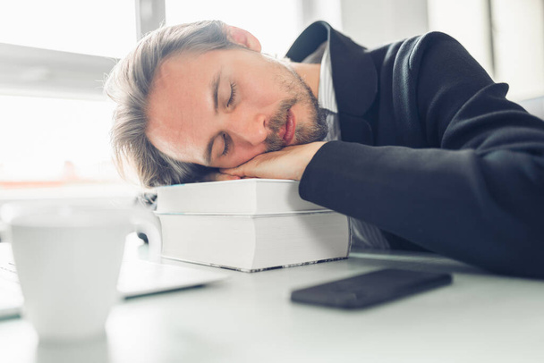 Handsome young man sleeping on pile of books at work desk. Coffe - Photo, image