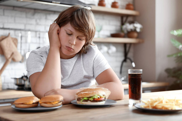 Emotional overweight boy at table with fast food in kitchen - Foto, Imagen