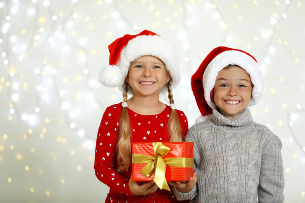 Happy little children in Santa hats with gift box against blurred festive lights. Christmas celebration - Фото, зображення