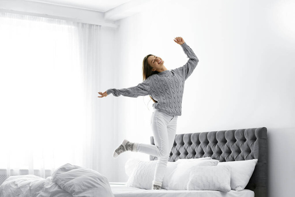Young woman in warm sweater jumping on bed at home - Foto, imagen