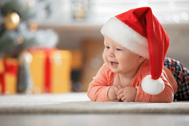 Little baby wearing Santa hat on floor indoors. First Christmas - Foto, imagen