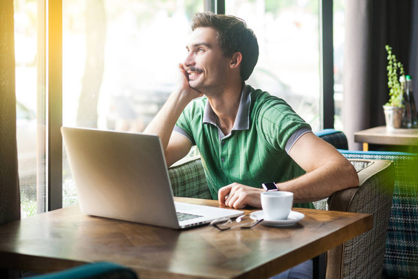 Young smiley happy businessman in green t-shirt looking outside while sitting at table with laptop in cafe, business and freelancing concept - Foto, afbeelding