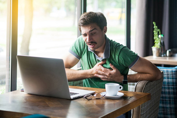 Young businessman in green t-shirt holding his chest and feeling pain on heart while sitting at table with laptop in cafe, business and freelancing concept - 写真・画像