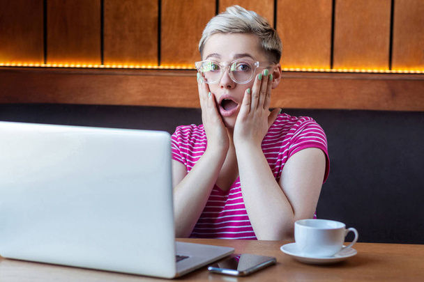 Wow! Portrait of emotional shocked young businesswoman in pink t-shirt is sitting in cafe, remotely working with suprised big eyes, open mouth and holding hands on face looking at camera.Indoor - Photo, Image