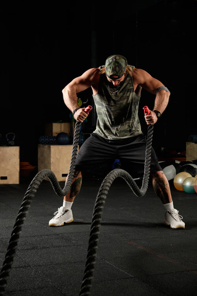 Muscular man in a sports uniform and a baseball cap stands with his head bowed with ropes in his hands in the gym - Foto, Imagem