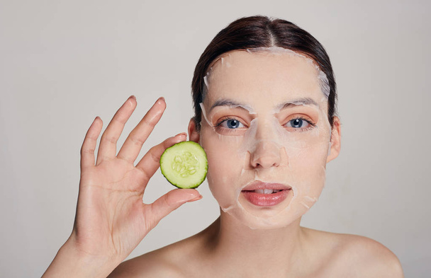 Close up gentle sophisticated calm girl in a moisturizing mask with a fresh cucumber on the face in the background serious with open eyes and right hand holds a cucumber near face - Foto, Bild