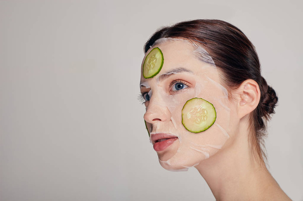 Zavřít jemný sofistikované dívka v hydratační masku s čerstvou okurkou na tváři v pozadí dívá pryč - Fotografie, Obrázek