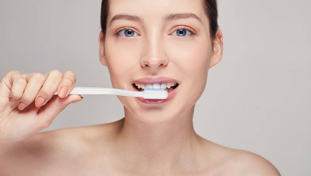 Young pretty girl with a toothbrush in her hands nearly her mouth with white teeth, with brown hair with clean fresh skin posing on a gray studio background - 写真・画像