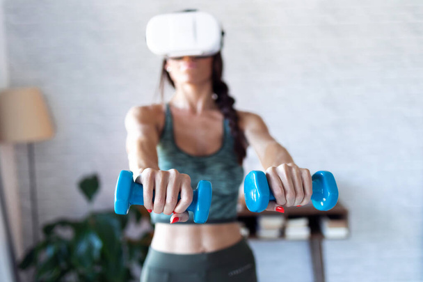 Beautiful young woman doing a workout with dumbbells with virtual reality headset at home. - Foto, Bild