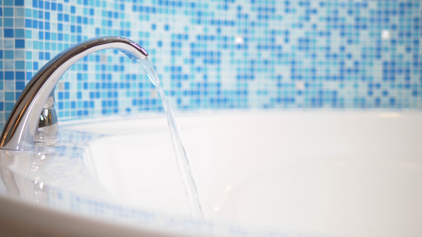 Nahaufnahme. Wasser fließt aus einem luxuriösen Wasserhahn in eine große moderne Badewanne vor einer blauen Mosaikwand - Filmmaterial, Video
