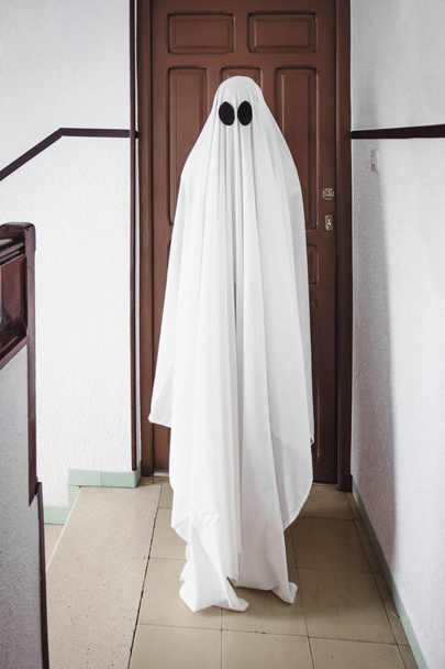 Geist bedeckt mit einem weißen Geistertuch vor einer alten Tür. Halloween-Konzept - Foto, Bild