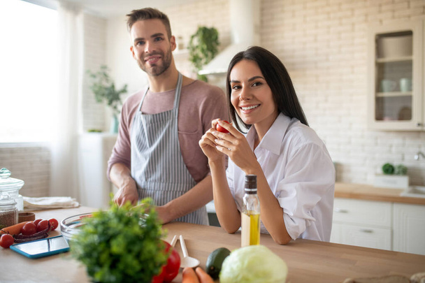 Dark-eyed wife holding tomato while cooking with husband - Photo, Image