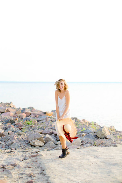 Pretty blonde woman walking on rocky beach with hant in hands and wearing dress. - Foto, afbeelding