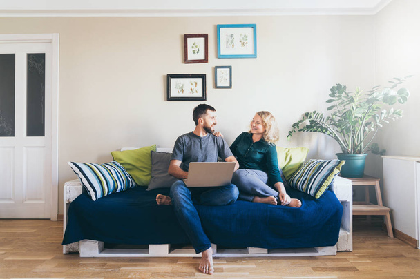 Młoda para robi zakupy online na kanapie w domu z laptopem, patrząc na siebie i uśmiechając się - Zdjęcie, obraz
