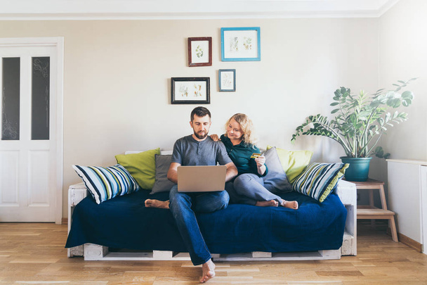 Casal jovem fazendo compras on-line no sofá em casa, olhando para a tela do laptop e sorrindo
 - Foto, Imagem