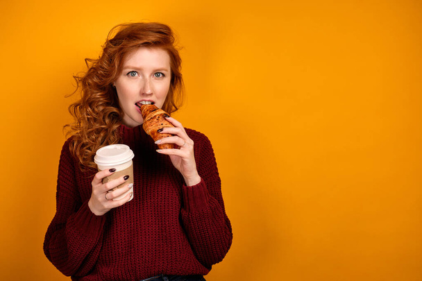 Red-haired curly girl in a burgundy sweater stands with the edge on a yellow background with a paper cup and bites a croissant. - Fotografie, Obrázek