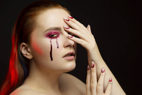 Scary beauty Halloween makeup portrait of a beautiful red hair girl with red eye shadows and fake blood from yellow eyes, studio portrait, red light, black background - Foto, imagen