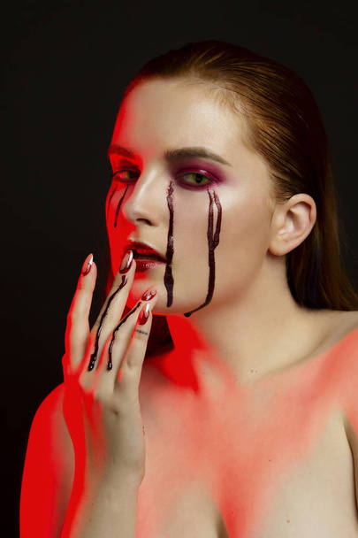 Scary beauty Halloween makeup portrait of a beautiful red hair girl with red eye shadows and fake blood from yellow eyes, studio portrait, red light, black background - Fotografie, Obrázek
