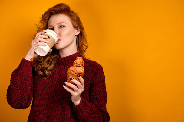 Red-haired curly girl in a burgundy sweater stands on a yellow background, takes a sip from a paper cup and holding a croissant. - Foto, afbeelding