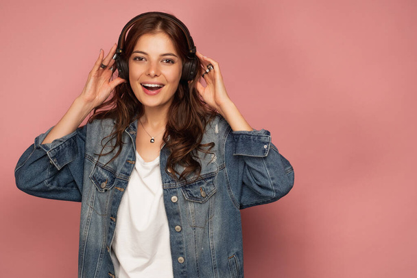 A dark-haired girl in jeans stands on a pink background laughing at the camera and holding headphones on her head - Fotó, kép