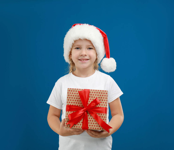 Cute little child wearing Santa hat with Christmas gift on blue background - Φωτογραφία, εικόνα