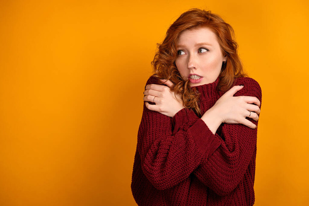 Beautiful red-haired girl in a burgundy sweater looks away clasping her shoulders with her hands. - Foto, Bild