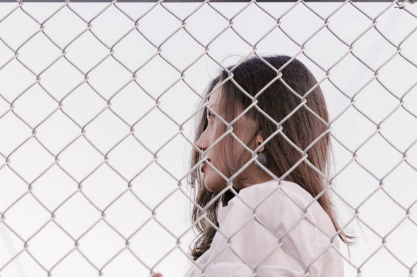 Pretty woman stands against wired fence. Model in white shirt. Purple lips. Pretty make up. Posing for magazine. Street fashion. Urban lifestyle. lady with long hair, beautiful eyes, behind fence. - Photo, Image