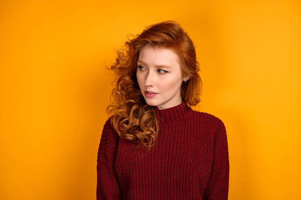 A red-haired girl in a burgundy sweater stands on a yellow background and looks to the side. - Foto, imagen