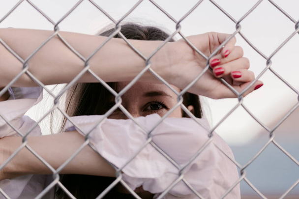 Pretty woman stands against wired fence. Model in white shirt. Purple lips. Pretty make up. Posing for magazine. Street fashion. Urban lifestyle. lady with long hair, beautiful eyes, behind fence. - Photo, Image