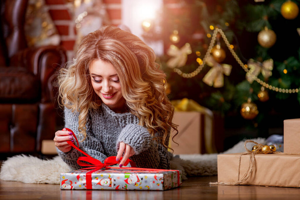 Girl in a sweater near the Christmas tree is unpacking a gift, New Year Christmas tree. Festive decorated New Year's room. Young woman in anticipation of Christmas. Blonde in grey knitted longsleeve. - 写真・画像