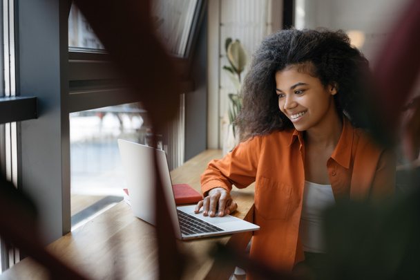 Güzel bir Afro-Amerikalı kadın ofiste başarılı olmak için çevrimiçi eğitim kursları izliyor. Dizüstü bilgisayar ve internet kullanan mutlu tasarımcının portresi, serbest çalışma projesi  - Fotoğraf, Görsel