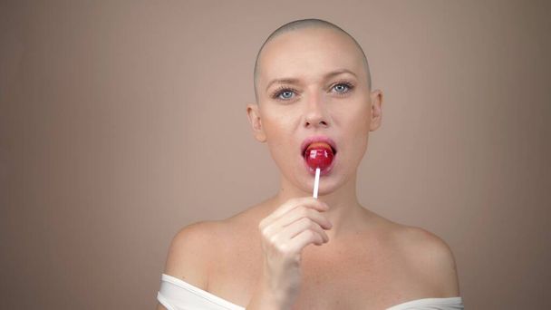 Beautiful bald girl eating a red round lollipop. beige background, copy space - Foto, Imagen