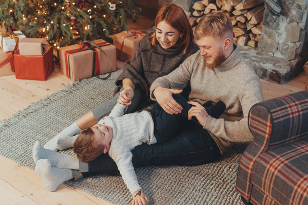 Νεαρή καυκάσια οικογένεια μαμά μπαμπάς γιος κοντά στο τζάκι χριστουγεννιάτικο δέντρο - Φωτογραφία, εικόνα