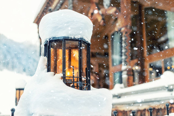 Acogedora lámpara de calle vintage con vidrio naranja cubierto de nieve y casa rústica de madera en el fondo al aire libre en las nevadas. Decoración de linterna de metal retro en la calle resort alpino. Navidad ambiente escena
 - Foto, Imagen
