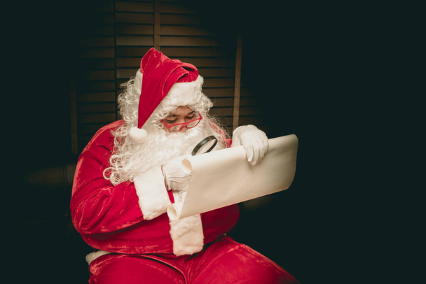 Père Noël utilisant une loupe pour regarder l'adresse des enfants pour apporter des cadeaux à envoyer ce soir
. - Photo, image