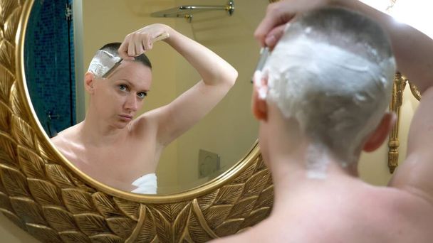 mooie kale vrouw kijkt in een luxe badkamer spiegel en doet haar haar, scheert haar haar met een gevaarlijk scheermes - Foto, afbeelding
