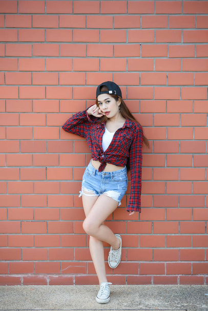 Chica hipsters asiáticos en la pared de ladrillo naranja, estilo de vida de la adolescencia moderna, dama feliz posando para tomar una foto, tailandia personas con una camisa a cuadros y pantalones cortos jeans
. - Foto, imagen