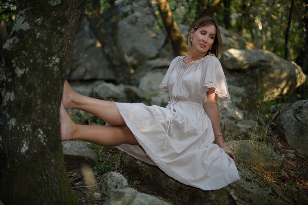 Nainen metsässä istuu ja rentouttaa jalkansa nojaten puuhun
 - Valokuva, kuva